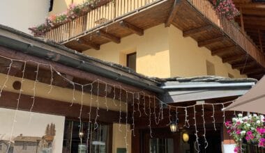 Charming alpine chalet with wooden balconies decorated with pink and white flowers, set against a bright blue sky, creating a picturesque mountain holiday retreat.